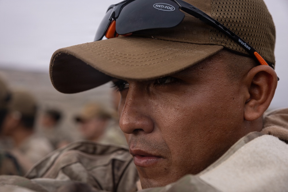 A close-up portrait of a male individual wearing a tan/olive baseball cap with anti-fog safety glasses pushed up on the brim. He is dressed in camouflage military-style clothing and appears focused or attentive. Several other individuals in similar attire are visible but blurred in the background.