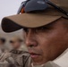 A close-up portrait of a male individual wearing a tan/olive baseball cap with anti-fog safety glasses pushed up on the brim. He is dressed in camouflage military-style clothing and appears focused or attentive. Several other individuals in similar attire are visible but blurred in the background.