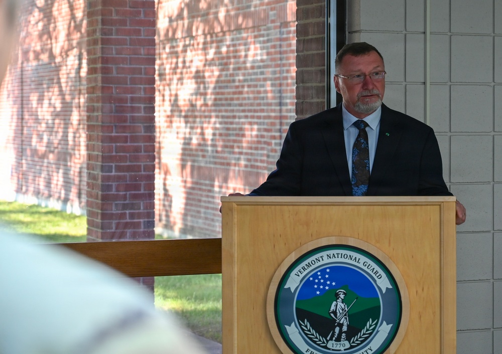 A man in a dark suit and tie is speaking at a podium bearing the Vermont National Guard seal. The setting appears to be an outdoor or semi-covered ceremonial event at a brick building. The speaker appears to be a civilian official or dignitary addressing an audience.