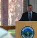 A man in a dark suit and tie is speaking at a podium bearing the Vermont National Guard seal. The setting appears to be an outdoor or semi-covered ceremonial event at a brick building. The speaker appears to be a civilian official or dignitary addressing an audience.