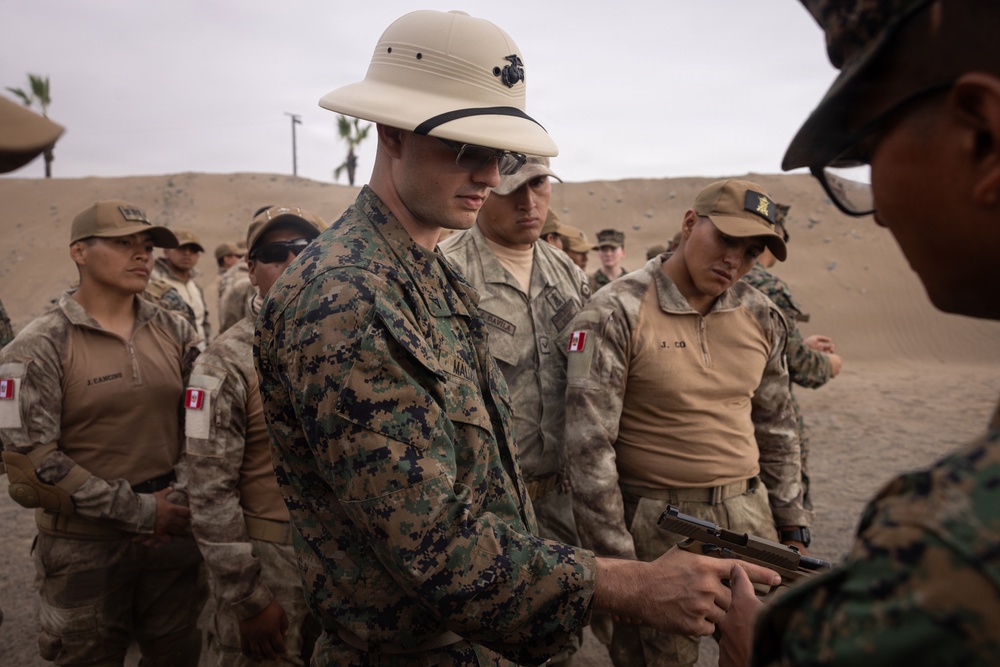 A group of military personnel from at least two different nations are gathered at what appears to be an outdoor firing range or training area in a desert environment. A U.S. Marine in MARPAT camouflage and a pith helmet is handling a pistol, likely during a weapons familiarization or training exercise. Several soldiers wearing Peruvian flag patches on their uniforms are observing, suggesting a joint military training exercise.