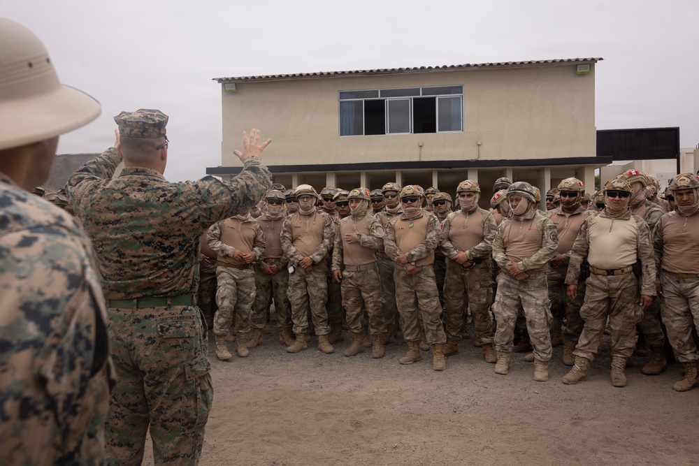 A military briefing or address is taking place outdoors in front of a two-story building. A figure in US Marine Corps digital camouflage uniform is gesturing with raised hand while speaking to a large formation of soldiers wearing tan/desert camouflage uniforms, helmets, face coverings, and eye protection. The assembled troops appear to be standing at ease in a loose formation, with at least one soldier wearing a red cross medical insignia.