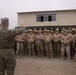A military briefing or address is taking place outdoors in front of a two-story building. A figure in US Marine Corps digital camouflage uniform is gesturing with raised hand while speaking to a large formation of soldiers wearing tan/desert camouflage uniforms, helmets, face coverings, and eye protection. The assembled troops appear to be standing at ease in a loose formation, with at least one soldier wearing a red cross medical insignia.