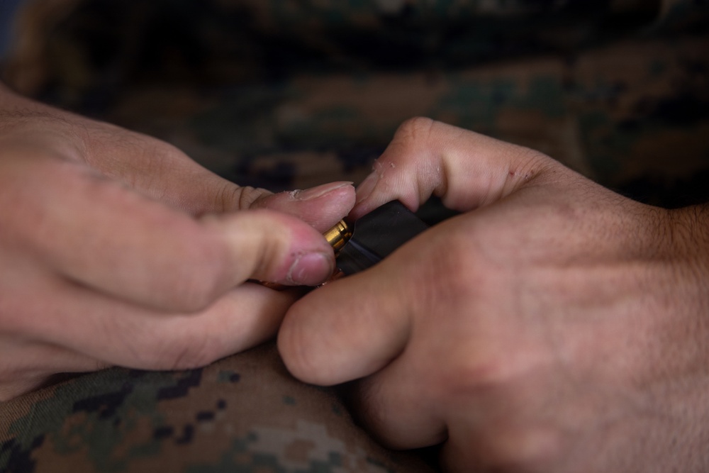 Close-up photograph of hands loading a ammunition round into a black firearm magazine. The person is wearing military camouflage (MARPAT pattern) uniform, indicating U.S. Marine Corps affiliation. The image is tightly focused on the hands and magazine, with the camouflage fabric visible in the background and foreground.