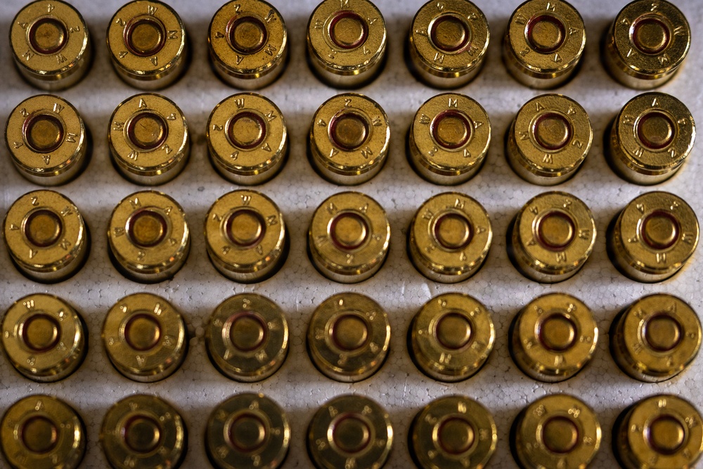 The image shows a close-up, top-down view of multiple brass ammunition cartridges arranged in uniform rows on a white/gray surface. The cartridges are viewed from the primer end (base), revealing headstamp markings on each round. The arrangement appears to be a standard factory ammunition tray or box layout.