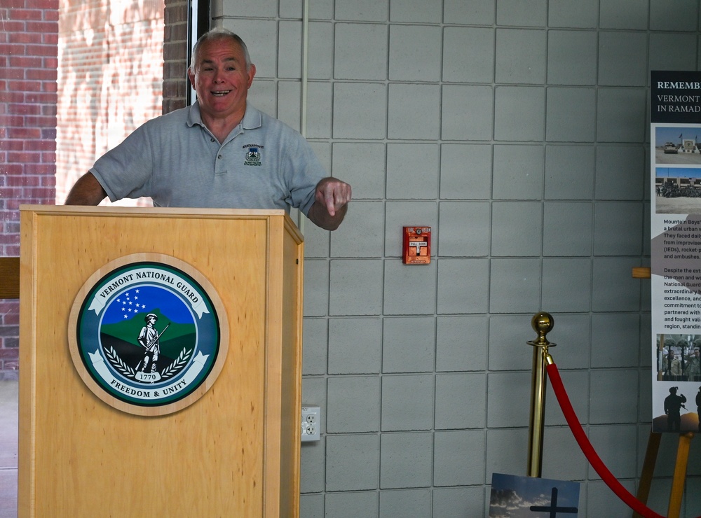A man in a gray polo shirt is speaking at a podium bearing the Vermont National Guard seal, which reads 'Freedom &amp; Unity' and '1770'. The setting appears to be an indoor ceremonial or commemorative event, with a display panel visible on the right referencing Vermont National Guard service in Ramadi. A red velvet rope barrier and photographs are visible in the background.