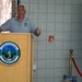 A man in a gray polo shirt is speaking at a podium bearing the Vermont National Guard seal, which reads 'Freedom &amp; Unity' and '1770'. The setting appears to be an indoor ceremonial or commemorative event, with a display panel visible on the right referencing Vermont National Guard service in Ramadi. A red velvet rope barrier and photographs are visible in the background.