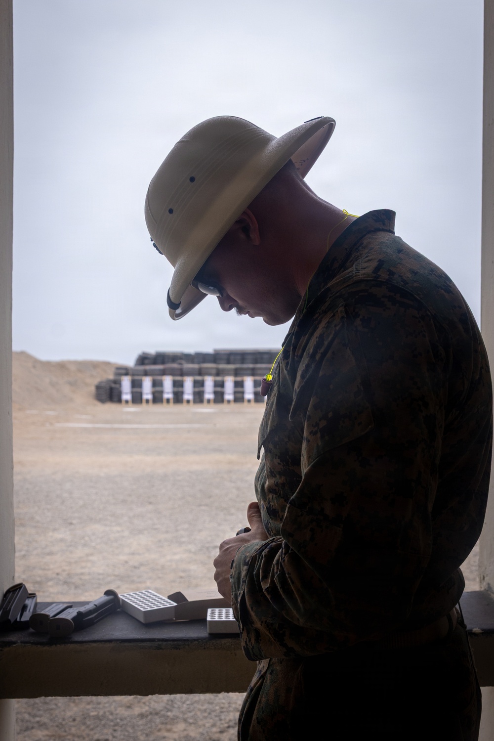 A military service member in MARPAT camouflage uniform and a tan drill instructor-style campaign cover (pith helmet) stands at a firing line station at an outdoor shooting range. The individual appears to be handling or loading ammunition at a shooting bench. Multiple paper targets on stands are visible downrange against a sandy berm backdrop.
