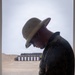 A military service member in MARPAT camouflage uniform and a tan drill instructor-style campaign cover (pith helmet) stands at a firing line station at an outdoor shooting range. The individual appears to be handling or loading ammunition at a shooting bench. Multiple paper targets on stands are visible downrange against a sandy berm backdrop.