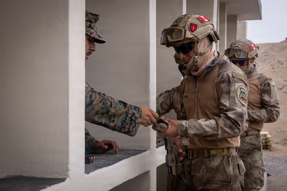Military personnel are engaged in what appears to be a weapons handling or ammunition exchange at a shooting range or training facility. A soldier in full tactical gear including helmet with red cross insignia, goggles, and face covering is receiving or handing over a magazine to a soldier in digital camouflage uniform. A third soldier in similar tactical gear is visible in the background.