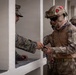 Military personnel are engaged in what appears to be a weapons handling or ammunition exchange at a shooting range or training facility. A soldier in full tactical gear including helmet with red cross insignia, goggles, and face covering is receiving or handing over a magazine to a soldier in digital camouflage uniform. A third soldier in similar tactical gear is visible in the background.