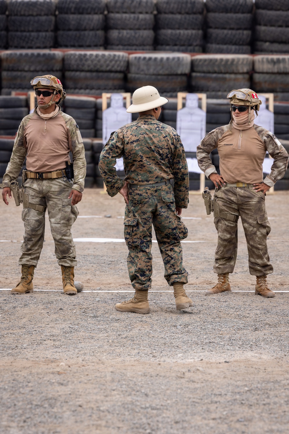 Three military personnel are standing at what appears to be an outdoor shooting range, with paper silhouette targets mounted on wooden frames visible in the background. Two soldiers wearing tan/desert camouflage uniforms with helmet-mounted goggles and flag patches face forward, while a third in woodland MARPAT camouflage and a pith helmet stands with their back to the camera. The setting includes a wall of stacked tires commonly used as a backstop at firing ranges.