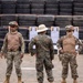 Three military personnel are standing at what appears to be an outdoor shooting range, with paper silhouette targets mounted on wooden frames visible in the background. Two soldiers wearing tan/desert camouflage uniforms with helmet-mounted goggles and flag patches face forward, while a third in woodland MARPAT camouflage and a pith helmet stands with their back to the camera. The setting includes a wall of stacked tires commonly used as a backstop at firing ranges.