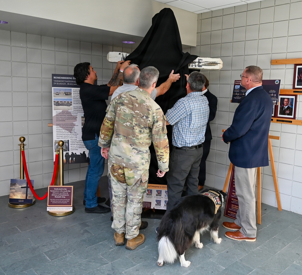 A group of individuals, including a uniformed military soldier, civilians in business and casual attire, are unveiling a memorial display by removing a black cloth covering. The display appears to be dedicated to Vermont National Guard members who served in Ramadi, Iraq. A service dog wearing a vest is present at the ceremony, and informational panels and a dedication plaque are visible nearby.