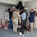 A group of individuals, including a uniformed military soldier, civilians in business and casual attire, are unveiling a memorial display by removing a black cloth covering. The display appears to be dedicated to Vermont National Guard members who served in Ramadi, Iraq. A service dog wearing a vest is present at the ceremony, and informational panels and a dedication plaque are visible nearby.