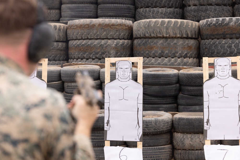 A military or law enforcement personnel in camouflage uniform is engaged in pistol shooting practice at a firing range. The backdrop consists of stacked used tires serving as a bullet stop, with paper humanoid silhouette targets mounted on wooden frames in the foreground. At least one target on the right shows visible bullet impact groupings in the chest area.