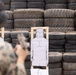 A military or law enforcement personnel in camouflage uniform is engaged in pistol shooting practice at a firing range. The backdrop consists of stacked used tires serving as a bullet stop, with paper humanoid silhouette targets mounted on wooden frames in the foreground. At least one target on the right shows visible bullet impact groupings in the chest area.