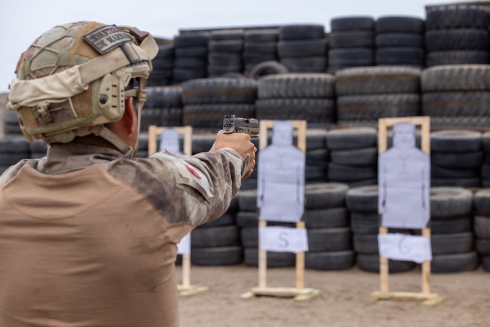 A military personnel member wearing desert camouflage and a tactical helmet with a patch reading 'Infantería de Marina' is aiming a pistol at paper silhouette targets on a shooting range. The targets, numbered 5 and 6, are mounted on wooden stands in front of a large wall of stacked tires serving as a backstop. This appears to be a marksmanship training exercise.