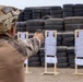 A military personnel member wearing desert camouflage and a tactical helmet with a patch reading 'Infantería de Marina' is aiming a pistol at paper silhouette targets on a shooting range. The targets, numbered 5 and 6, are mounted on wooden stands in front of a large wall of stacked tires serving as a backstop. This appears to be a marksmanship training exercise.