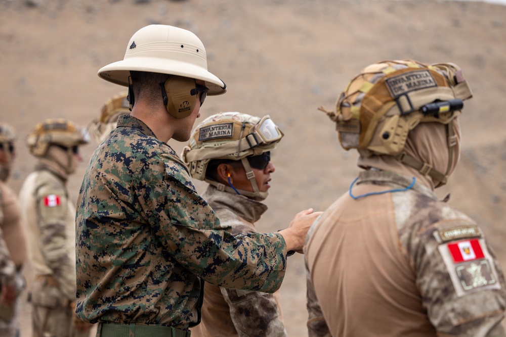 The image shows military personnel engaged in what appears to be a training or briefing exercise in an arid, desert-like environment. At least two soldiers wear helmets marked 'Infanteria de Marina' (Marine Infantry) with Peruvian flag patches, while one individual in green digital camouflage and a pith helmet appears to be instructing or inspecting them. The setting and insignia suggest a joint military exercise involving Peruvian Marine Infantry.