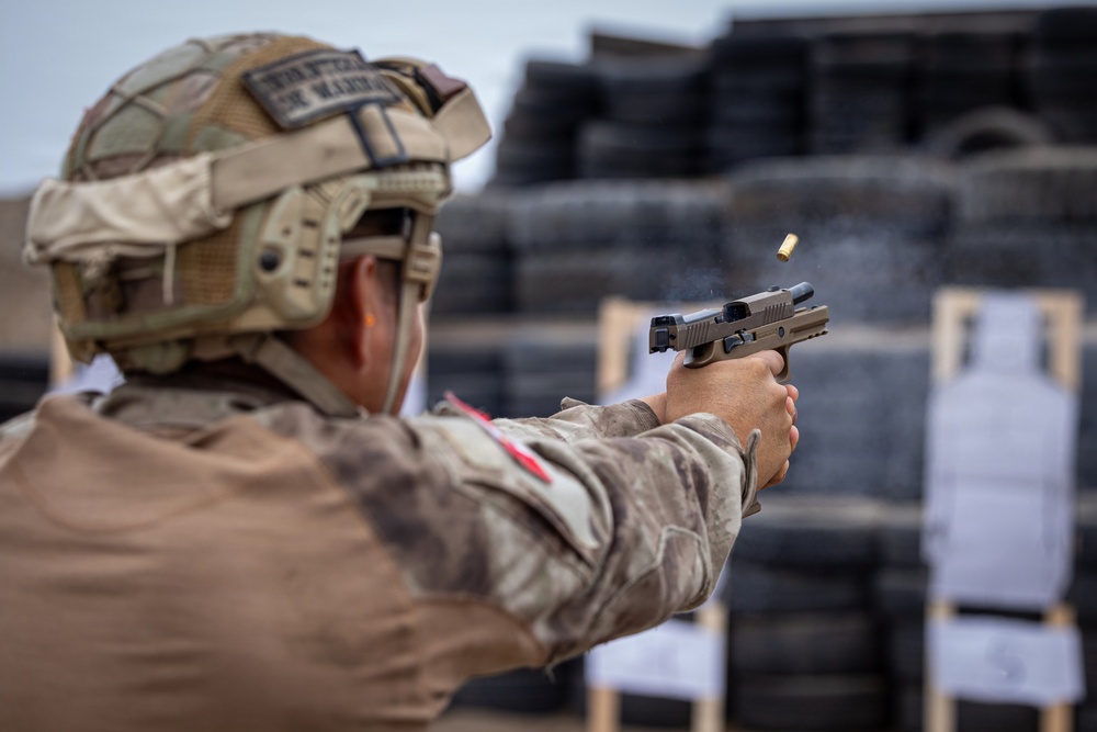 A military or law enforcement personnel in full tactical gear is firing a semi-automatic pistol at a shooting range. A spent brass casing is visibly ejecting from the firearm mid-shot. The background shows stacked tires used as a backstop and a paper target on a wooden stand.