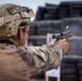 A military or law enforcement personnel in full tactical gear is firing a semi-automatic pistol at a shooting range. A spent brass casing is visibly ejecting from the firearm mid-shot. The background shows stacked tires used as a backstop and a paper target on a wooden stand.