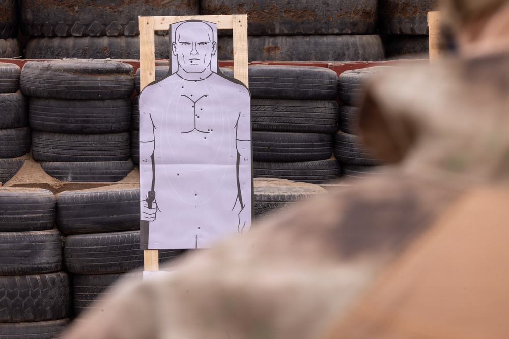 A shooting range training scenario showing a paper human silhouette target mounted on a wooden frame, backed by stacked tires used as a bullet stop. The target displays multiple bullet hole impacts concentrated in the chest/torso area. A person in camouflage military or tactical attire is visible in the foreground, blurred out of focus.
