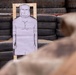 A shooting range training scenario showing a paper human silhouette target mounted on a wooden frame, backed by stacked tires used as a bullet stop. The target displays multiple bullet hole impacts concentrated in the chest/torso area. A person in camouflage military or tactical attire is visible in the foreground, blurred out of focus.