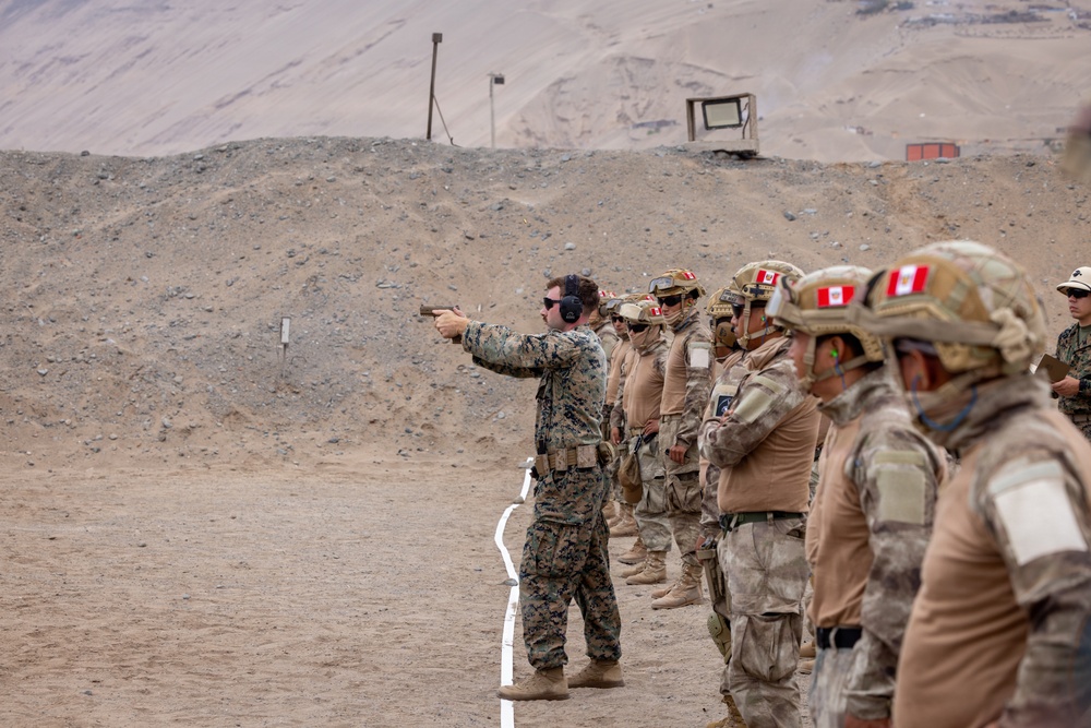 A military training exercise at an outdoor shooting range in an arid, desert-like environment. A soldier in US Marine Corps digital camouflage is demonstrating pistol shooting technique while a line of soldiers wearing tan/desert camouflage with red and white flag patches on their helmets observe. The setting appears to be a joint multinational military training exercise.