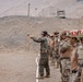 A military training exercise at an outdoor shooting range in an arid, desert-like environment. A soldier in US Marine Corps digital camouflage is demonstrating pistol shooting technique while a line of soldiers wearing tan/desert camouflage with red and white flag patches on their helmets observe. The setting appears to be a joint multinational military training exercise.