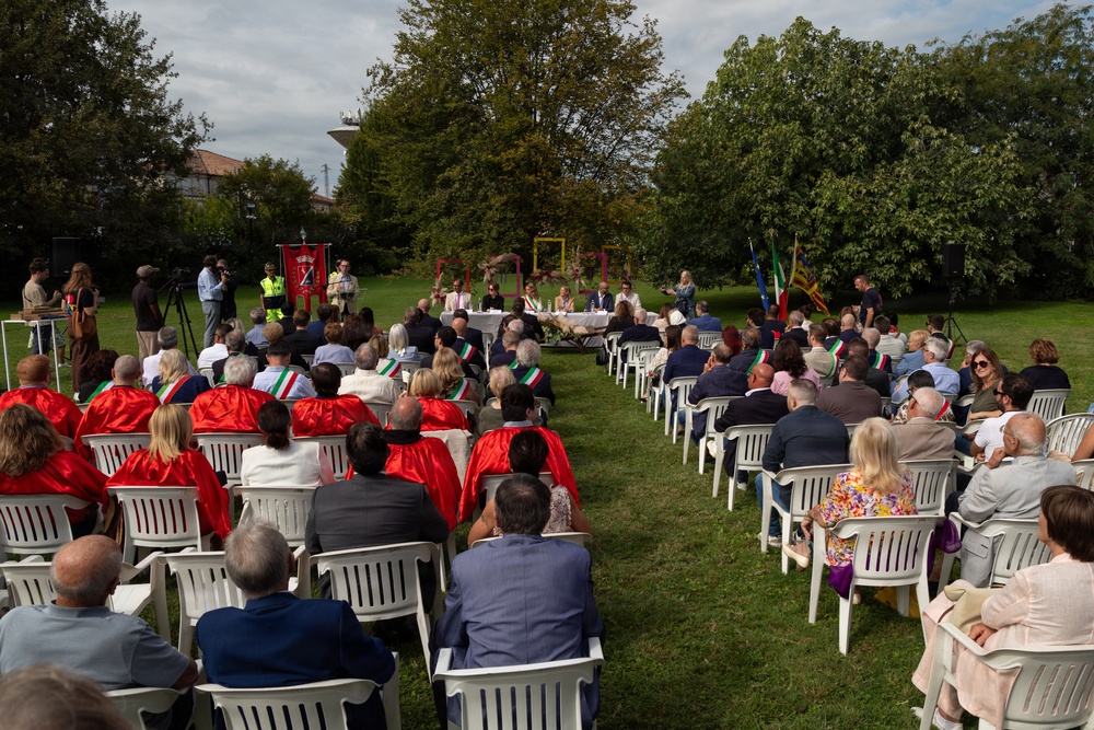 An outdoor public ceremony or civic event taking place on a grass lawn, with a large audience seated in white plastic chairs arranged in two sections facing a head table with officials. Several attendees wear red capes and Italian tricolor sashes, suggesting a formal municipal or civic occasion. Flags including what appears to be the Italian flag and EU flag are visible near the front, along with a ceremonial banner/standard.