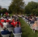 An outdoor public ceremony or civic event taking place on a grass lawn, with a large audience seated in white plastic chairs arranged in two sections facing a head table with officials. Several attendees wear red capes and Italian tricolor sashes, suggesting a formal municipal or civic occasion. Flags including what appears to be the Italian flag and EU flag are visible near the front, along with a ceremonial banner/standard.