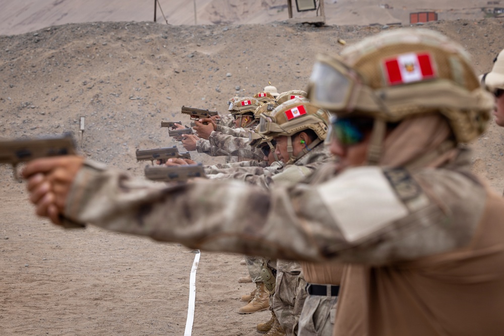 A line of military personnel in desert camouflage uniforms are conducting a pistol shooting exercise at an outdoor firing range. The soldiers are wearing tactical helmets bearing what appears to be the Peruvian flag patch, along with eye and ear protection. The setting is an arid, sandy environment consistent with a military training facility.