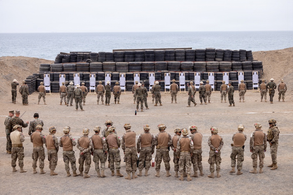 A military shooting range exercise is taking place in a coastal desert environment. Two groups of soldiers are visible: one group standing at firing positions facing paper targets mounted in front of a large tire backstop wall, and another group in the foreground being briefed by instructors. The setting appears to be a joint or multinational training exercise based on the mix of uniform styles and insignia.