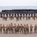 A military shooting range exercise is taking place in a coastal desert environment. Two groups of soldiers are visible: one group standing at firing positions facing paper targets mounted in front of a large tire backstop wall, and another group in the foreground being briefed by instructors. The setting appears to be a joint or multinational training exercise based on the mix of uniform styles and insignia.