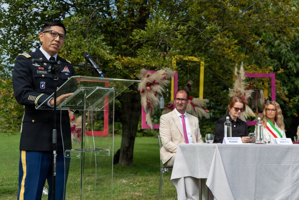 A U.S. Army officer in dress uniform is delivering a speech at an outdoor podium during what appears to be an official ceremony or commemorative event. Behind the speaker, three civilians are seated at a draped table with name placards visible, including 'Marcello Rosano' and what appears to be 'Rizzotto'. The setting is a well-maintained outdoor garden with decorative pink frame installations and floral arrangements.