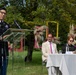 A U.S. Army officer in dress uniform is delivering a speech at an outdoor podium during what appears to be an official ceremony or commemorative event. Behind the speaker, three civilians are seated at a draped table with name placards visible, including 'Marcello Rosano' and what appears to be 'Rizzotto'. The setting is a well-maintained outdoor garden with decorative pink frame installations and floral arrangements.