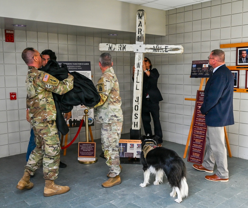 A memorial ceremony is taking place in what appears to be a military building, centered around a large white wooden cross bearing names including 'MARK,' 'MILE,' 'WILL,' and 'JOSH.' Several military personnel in camouflage uniforms, a civilian woman in black, and a man in a suit are gathered around the memorial display. A black-and-white border collie wearing a vest is present near the base of the cross.