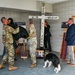 A memorial ceremony is taking place in what appears to be a military building, centered around a large white wooden cross bearing names including 'MARK,' 'MILE,' 'WILL,' and 'JOSH.' Several military personnel in camouflage uniforms, a civilian woman in black, and a man in a suit are gathered around the memorial display. A black-and-white border collie wearing a vest is present near the base of the cross.