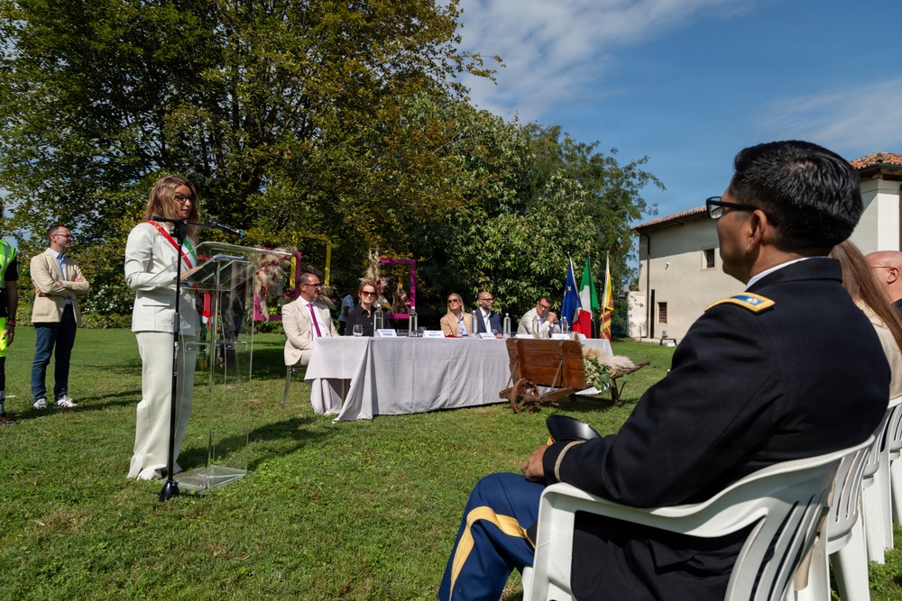 An outdoor official ceremony or civic event is taking place on a grass lawn, likely in Italy based on the visible Italian flag and architectural style. A woman in a white suit is delivering a speech at a transparent podium while dignitaries and officials are seated at a draped table behind her. A uniformed U.S. Army officer is seated in the foreground as part of the audience.