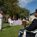 An outdoor official ceremony or civic event is taking place on a grass lawn, likely in Italy based on the visible Italian flag and architectural style. A woman in a white suit is delivering a speech at a transparent podium while dignitaries and officials are seated at a draped table behind her. A uniformed U.S. Army officer is seated in the foreground as part of the audience.