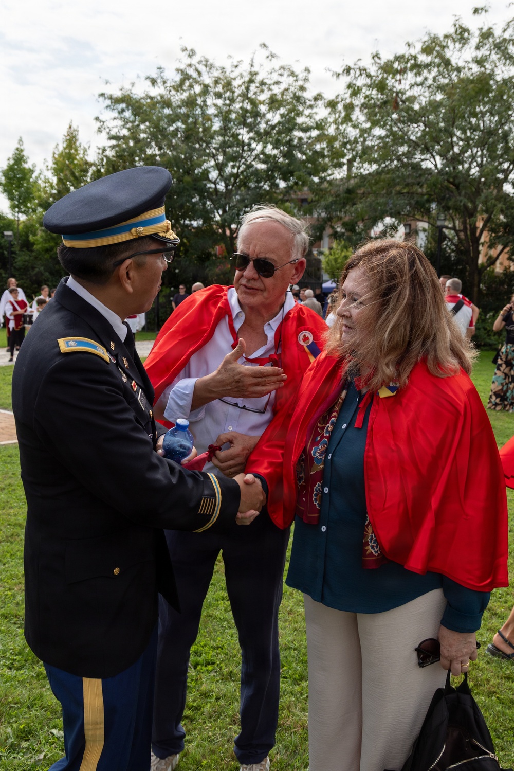 A U.S. Army officer in dress uniform is shaking hands and conversing with two civilians wearing red capes or cloaks at an outdoor event. The two civilians, a man and a woman, appear to be wearing Canadian-themed attire with maple leaf pins visible. The setting appears to be a formal or ceremonial outdoor gathering on a grassy area.