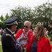 A U.S. Army officer in dress uniform is shaking hands and conversing with two civilians wearing red capes or cloaks at an outdoor event. The two civilians, a man and a woman, appear to be wearing Canadian-themed attire with maple leaf pins visible. The setting appears to be a formal or ceremonial outdoor gathering on a grassy area.