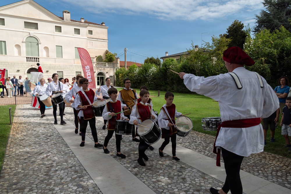 A traditional Italian historical parade or festival featuring children and adults dressed in medieval-style costumes of white and red/burgundy colors, marching while playing drums. A conductor or leader in period costume directs the procession with a baton. The event takes place in front of a classical Italian villa or palazzo, suggesting a local heritage celebration.