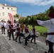 A traditional Italian historical parade or festival featuring children and adults dressed in medieval-style costumes of white and red/burgundy colors, marching while playing drums. A conductor or leader in period costume directs the procession with a baton. The event takes place in front of a classical Italian villa or palazzo, suggesting a local heritage celebration.