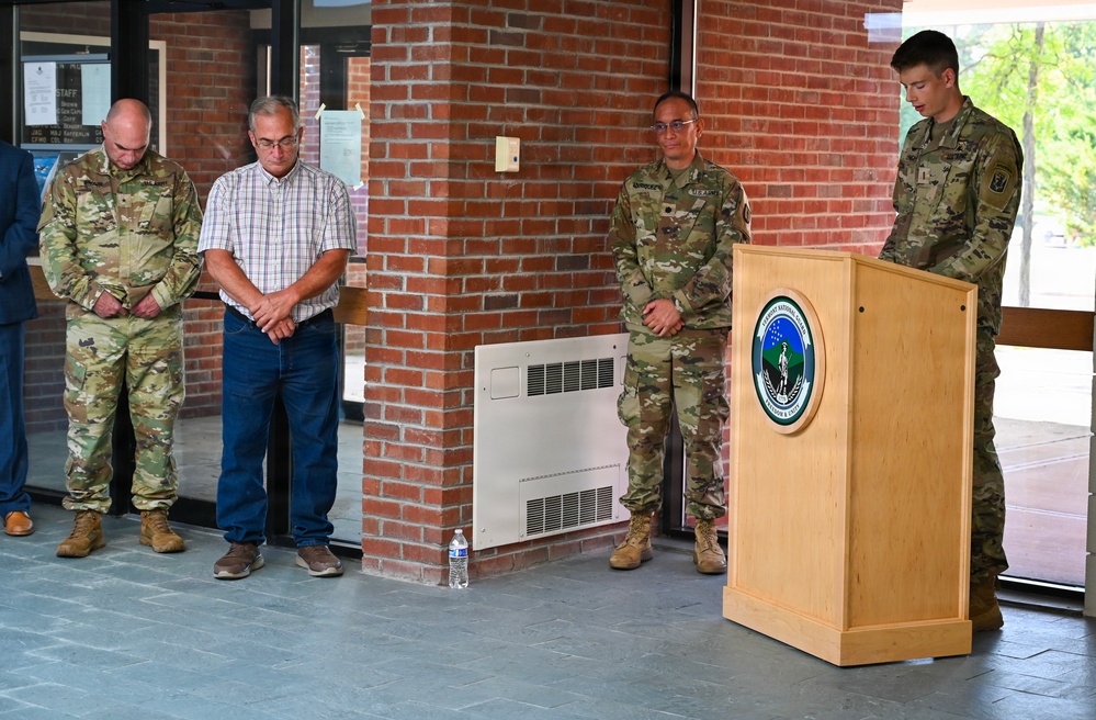 A formal military ceremony is taking place at what appears to be a U.S. Army installation, with personnel standing at a podium bearing an official seal. Several individuals, including uniformed soldiers in OCP (Operational Camouflage Pattern) uniforms and one civilian, have their heads bowed in what appears to be a moment of prayer or reflection. The setting is an outdoor covered entrance area of a brick building.