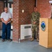 A formal military ceremony is taking place at what appears to be a U.S. Army installation, with personnel standing at a podium bearing an official seal. Several individuals, including uniformed soldiers in OCP (Operational Camouflage Pattern) uniforms and one civilian, have their heads bowed in what appears to be a moment of prayer or reflection. The setting is an outdoor covered entrance area of a brick building.