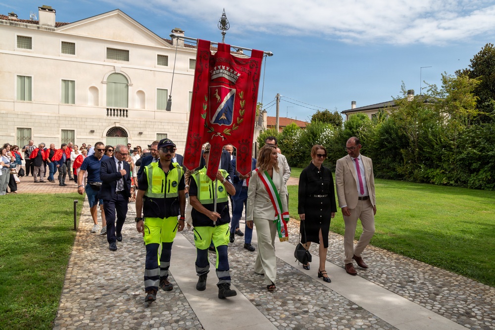 A formal civic procession is taking place outdoors, likely in an Italian municipality, led by two municipal police officers in high-visibility yellow uniforms carrying a large red ceremonial banner bearing a coat of arms and crown. Behind them walks a woman wearing the Italian tricolor mayoral sash (fascia tricolore), accompanied by other officials and a crowd of attendees. The setting appears to be the grounds of a historic villa or municipal building in Italy.