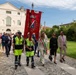 A formal civic procession is taking place outdoors, likely in an Italian municipality, led by two municipal police officers in high-visibility yellow uniforms carrying a large red ceremonial banner bearing a coat of arms and crown. Behind them walks a woman wearing the Italian tricolor mayoral sash (fascia tricolore), accompanied by other officials and a crowd of attendees. The setting appears to be the grounds of a historic villa or municipal building in Italy.