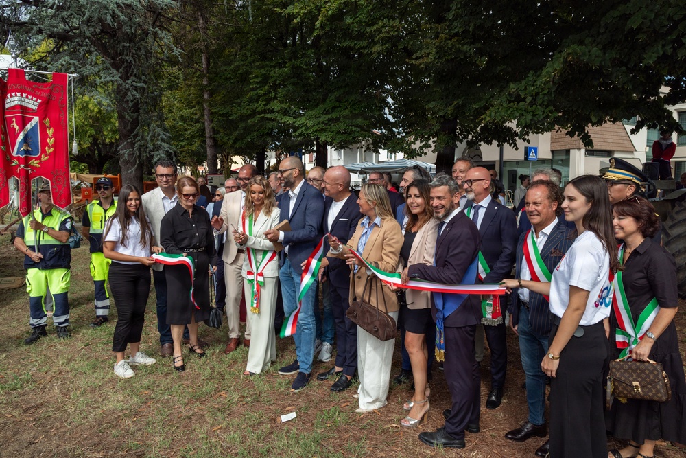 A formal ribbon-cutting ceremony is taking place outdoors in what appears to be an Italian municipality, with officials and dignitaries holding a ribbon in the Italian national colors (green, white, and red). Several participants are wearing ceremonial sashes also in Italian tricolor colors, indicating they are likely elected officials or mayors. A municipal banner/gonfalon is visible on the left side of the image.